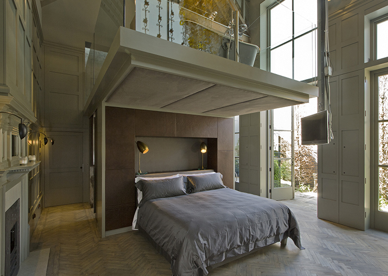 Master bedroom in the old coach house reuses the original parquet floor from the old workshop and has double height shuttered windows onto a walled garden. The TV can be winched up to the mezzanine to watch movies in the bath above.